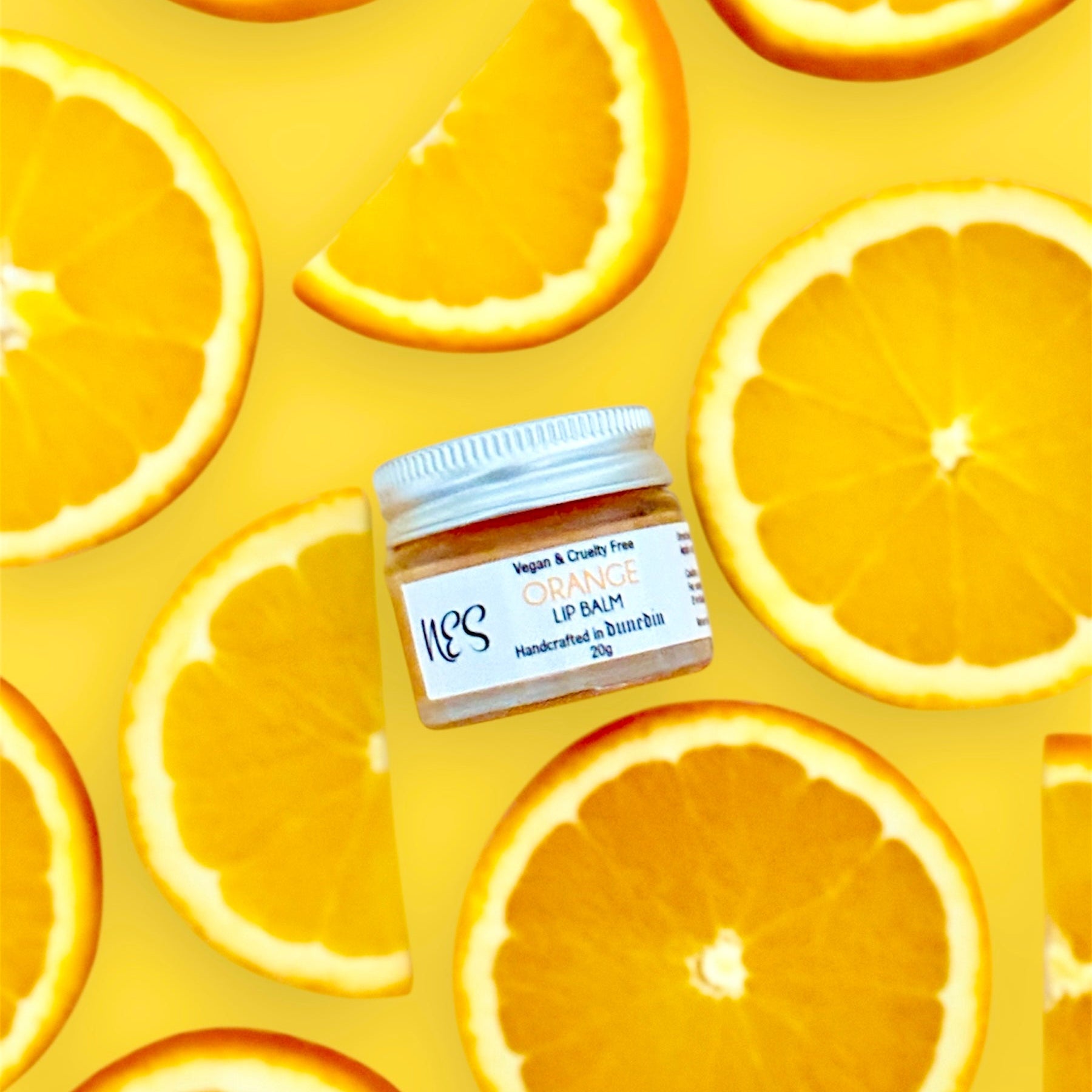 NES Orange Lip Balm in a glass jar, surrounded by vibrant orange slices on a yellow background. Vegan, cruelty-free, and handcrafted in Dunedin, NZ.