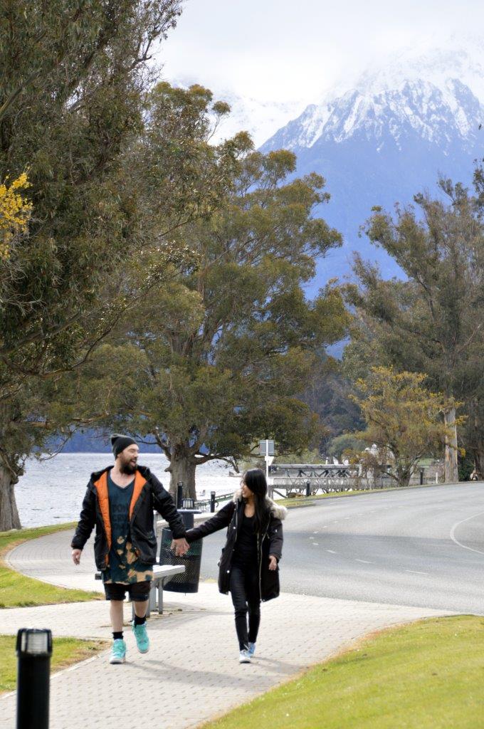 Elza and Nathaniel walking hand in hand along a lakeside path in New Zealand, with scenic mountains and trees in the background. Outdoor lifestyle in NZ.