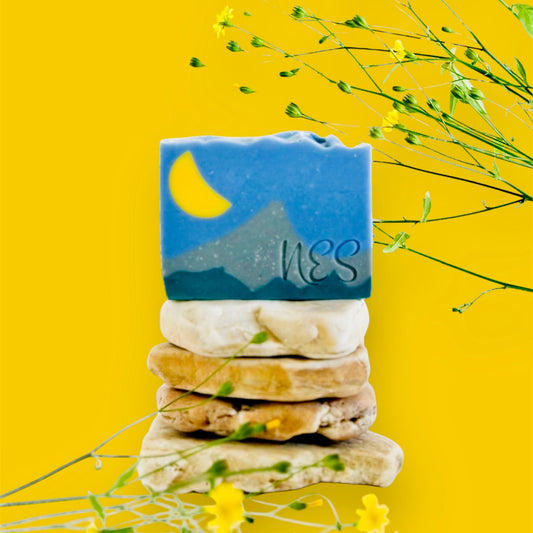 NES Soap’s Silver Peaks artisan soap, featuring a moonlit mountain design, stacked on natural stones with delicate yellow wildflowers in the background.